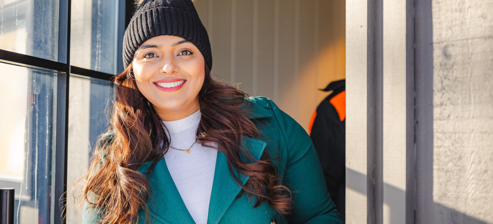 A woman dressed in a winter coat and hat smiles and stands at the entrance of a business.